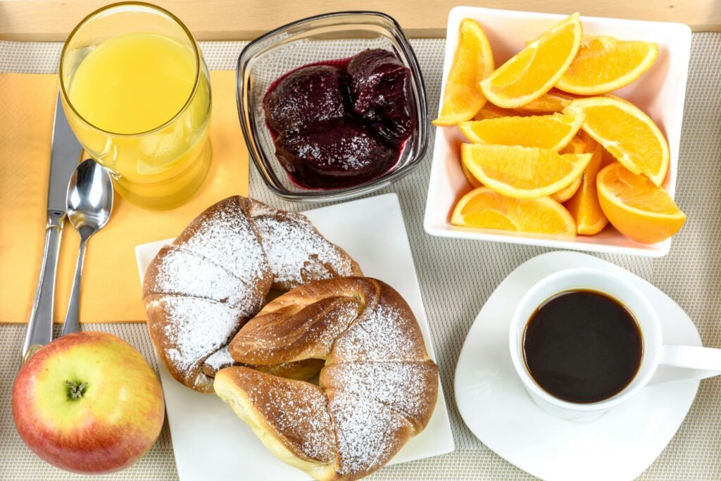 Fresh continental breakfast with croissant, coffee, orange slices, juice, jam, and apple, served on a plate.