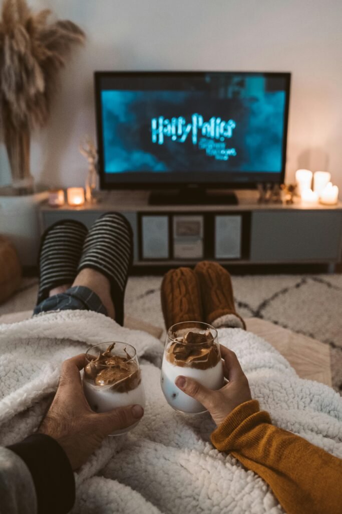 A couple enjoys a cozy movie night with drinks, wrapped in blankets, watching a Harry Potter film on TV.